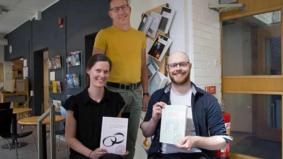 En grupp med tre personer, två av dem håller upp varsin publikation. Foto.